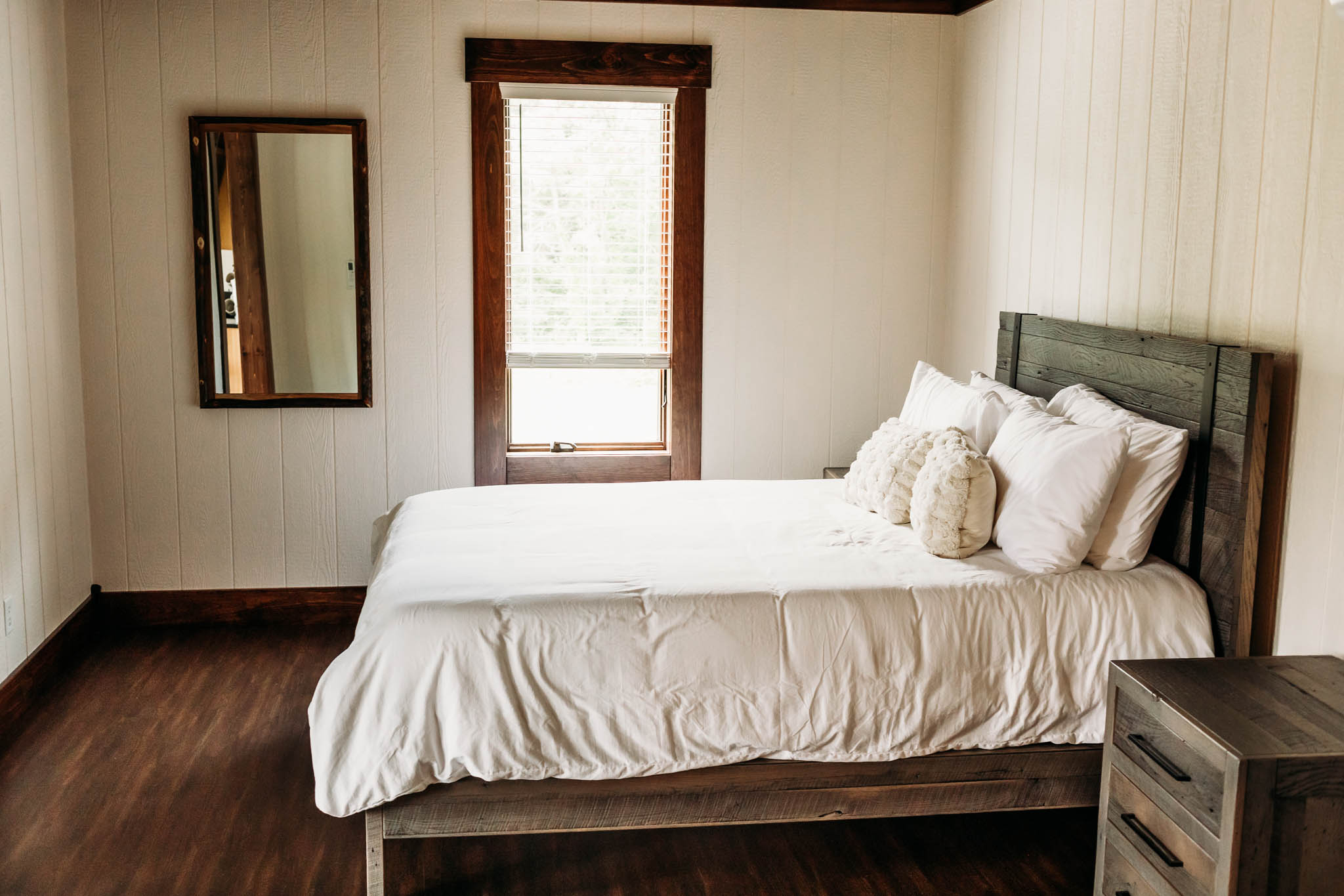 A view of the bedroom in the Villas at The VeNue. There is a wooden queen bed with white covers, wooden nightstand, large window and mirror hanging on the wall.