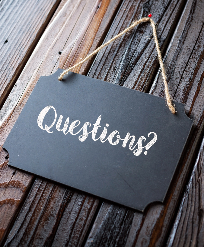 Rectangular chalk board sign with "Questions?" written in cursive white chalk. The chalk board is laying on dark wood boards.