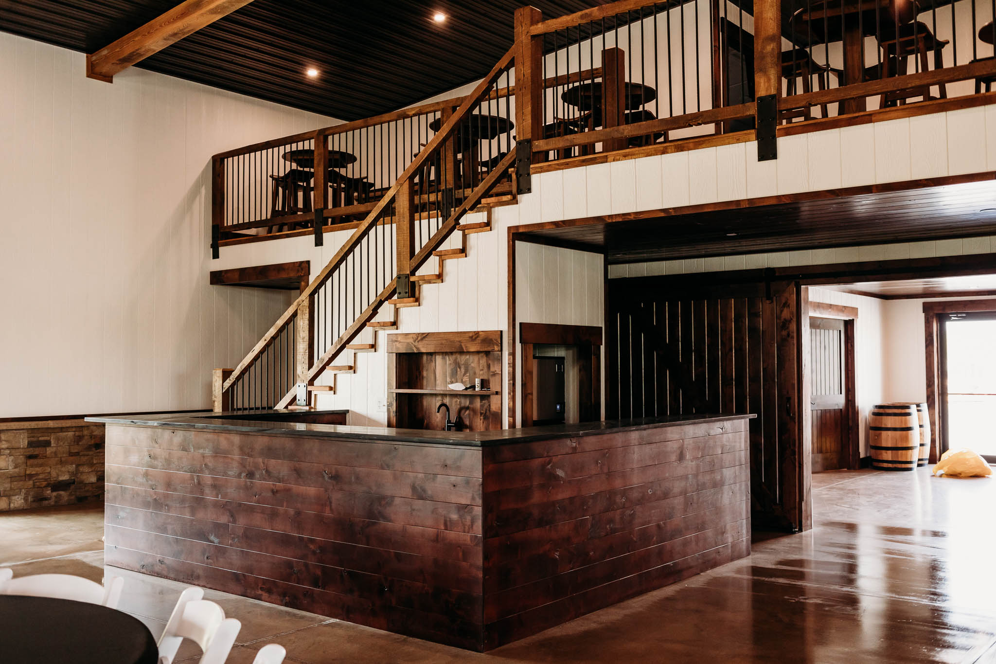 A wide view of the drink serving bar and stairs to the upper loft seating area. The bar is made of dark walnut wood and the handrails of the stairs are reclaimed oak wood.