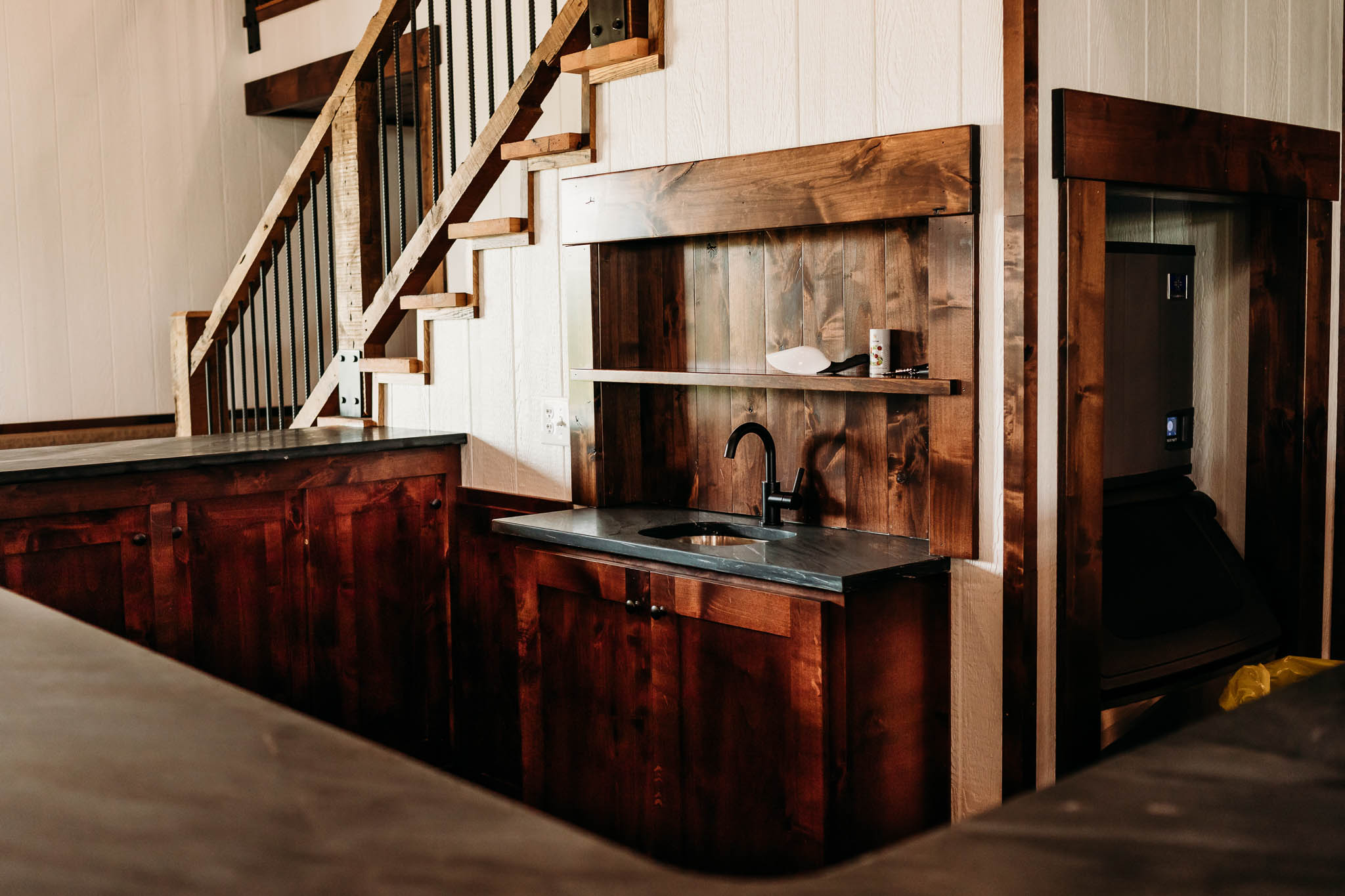 Drink serving bar in The VeNue Event Space with a small sink, and cabinets made of dark walnut wood are under the black granite counter top. An ice machine is in the corner on the right.
