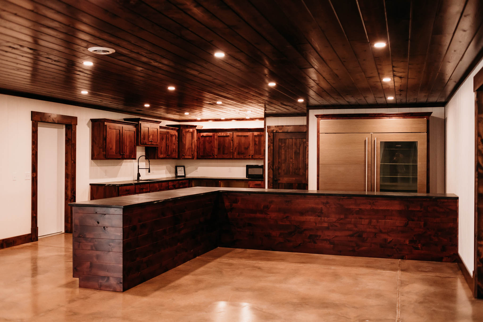 The caterer's kitchen in The VeNue Event Space. The kitchen cabinets are a dark walnut finish there is a large refrigerator on the right side and microwave, sink and warming drawers on the left side.