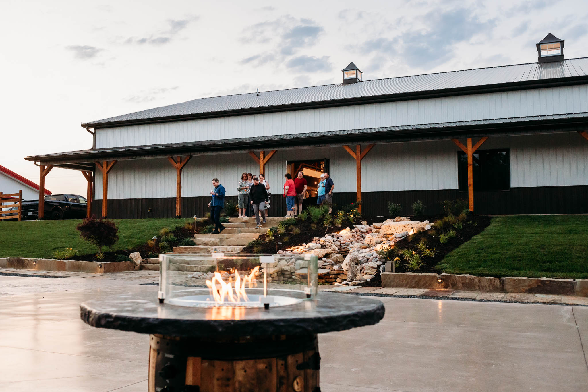 Backside view of The VeNue Event Space with people walking down the rock stairs and lit whiskey barrel firepit in the foreground