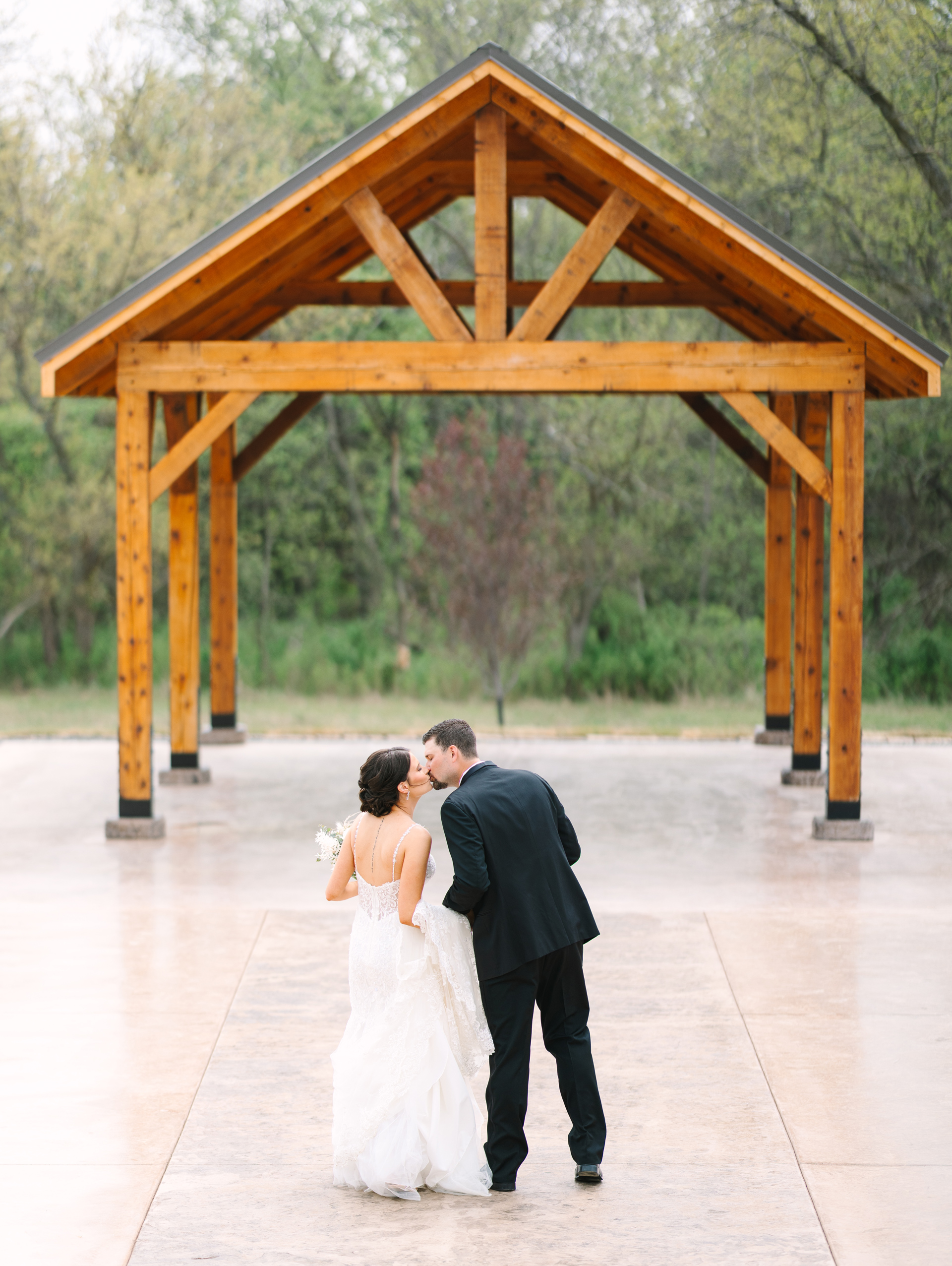A caucasian bride and groom stop to kiss in front of a wooden pavilion surrounded by trees