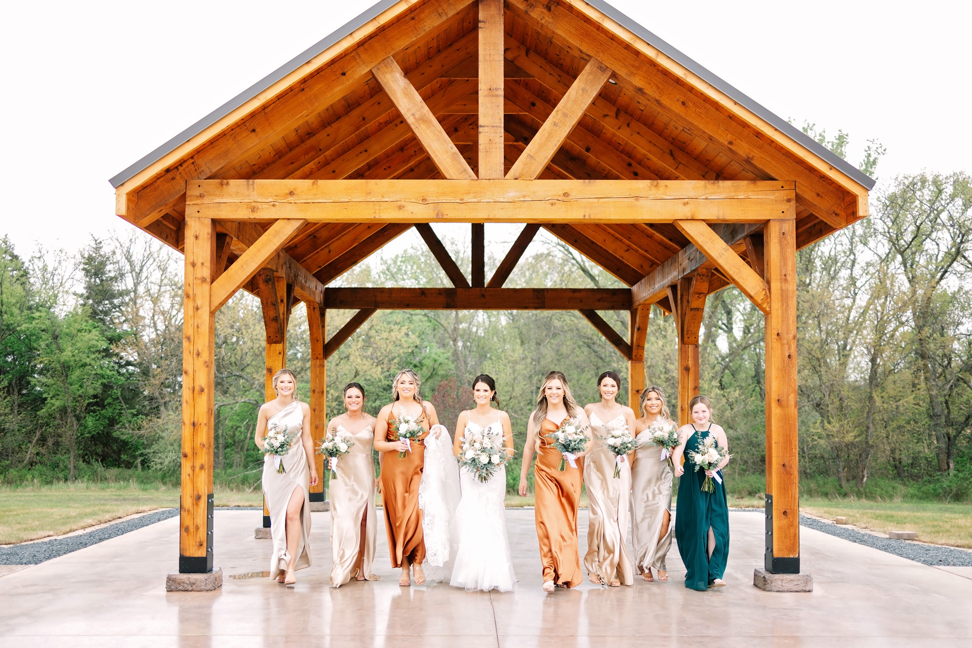 A Caucasian bride and 7 bridesmaids in cream, orange and green dresses in front of a wooden pavilion. Photography copyright Alicia Castañeda Photography.