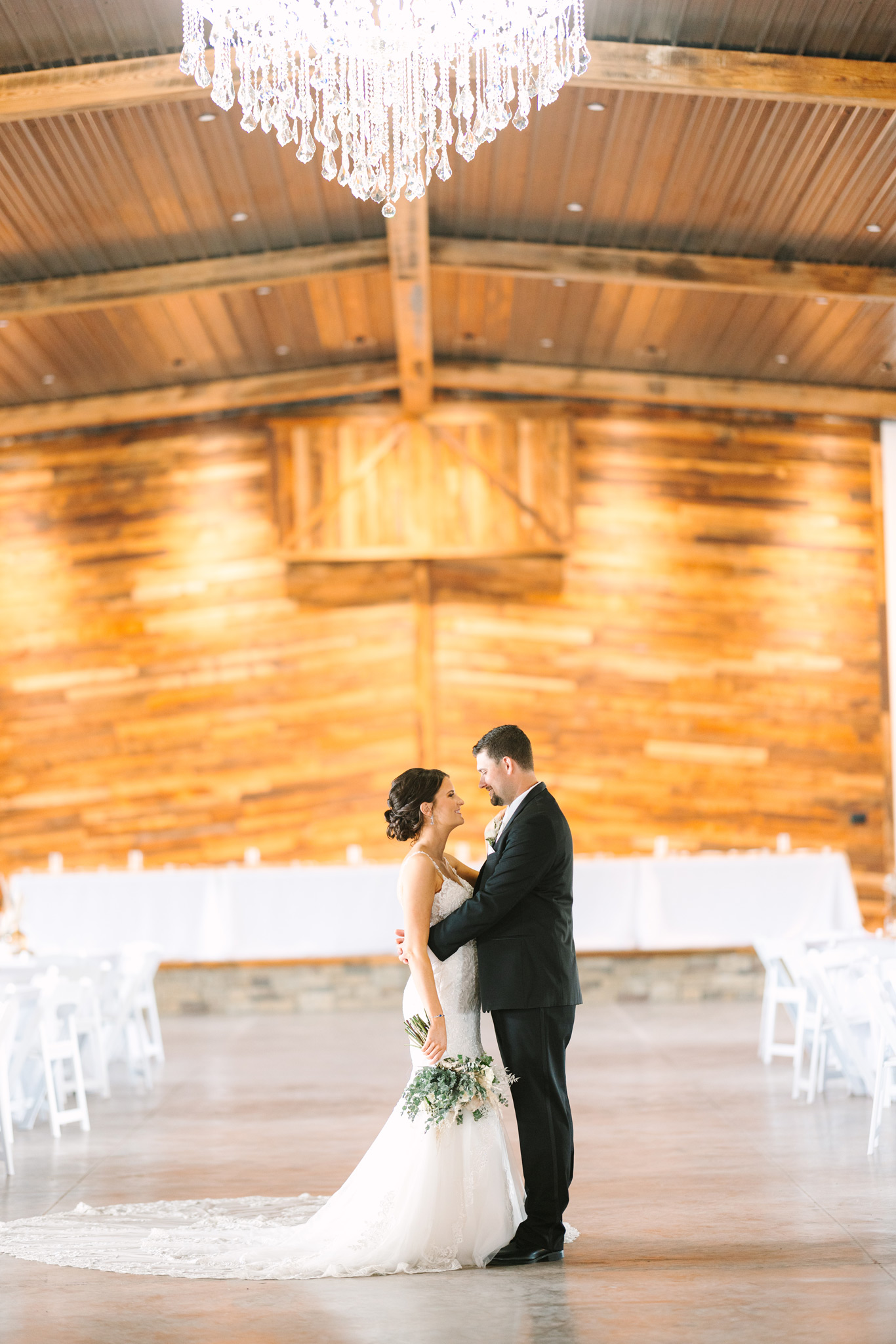 Bride and groom embrace each other in between white tables and chairs in front of a large wooden stage