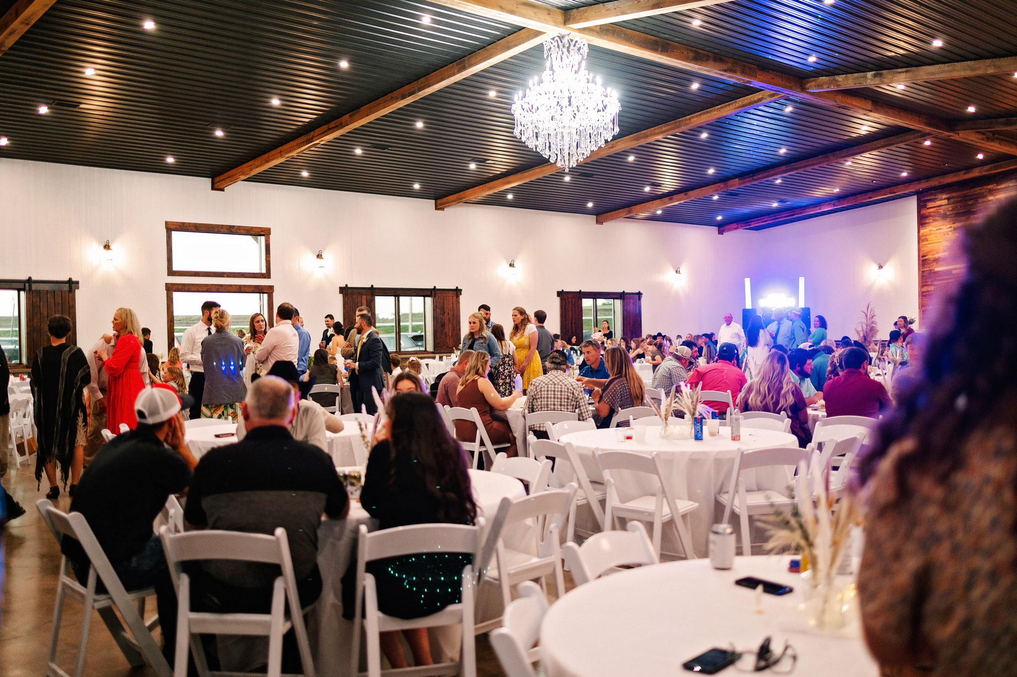 A large group of people sitting and standing at tables and chairs in white linens at The VeNue Event Space