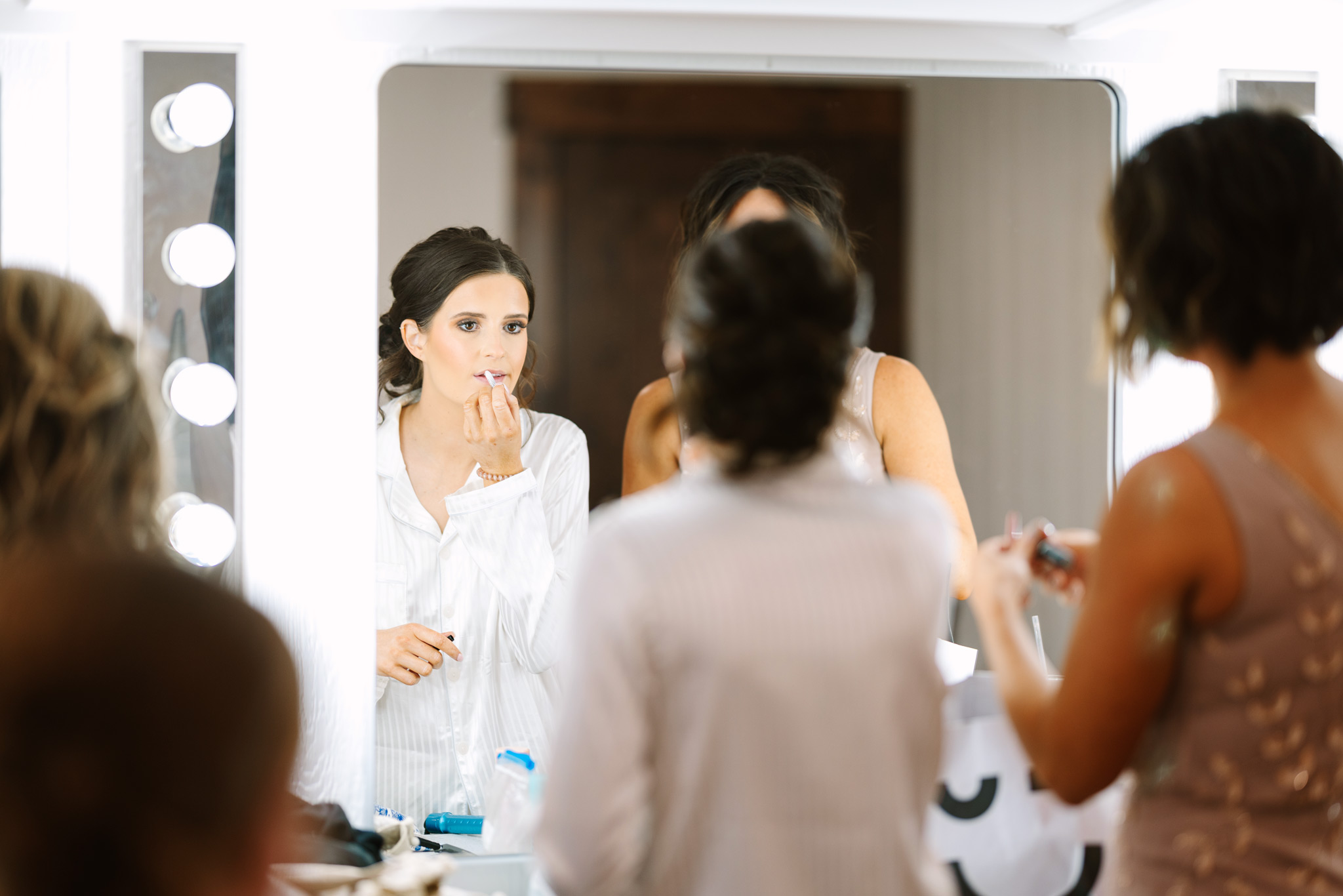 Bride with dark hair facing a large mirror with Hollywood lighting and putting on make up at The VeNue Event Space