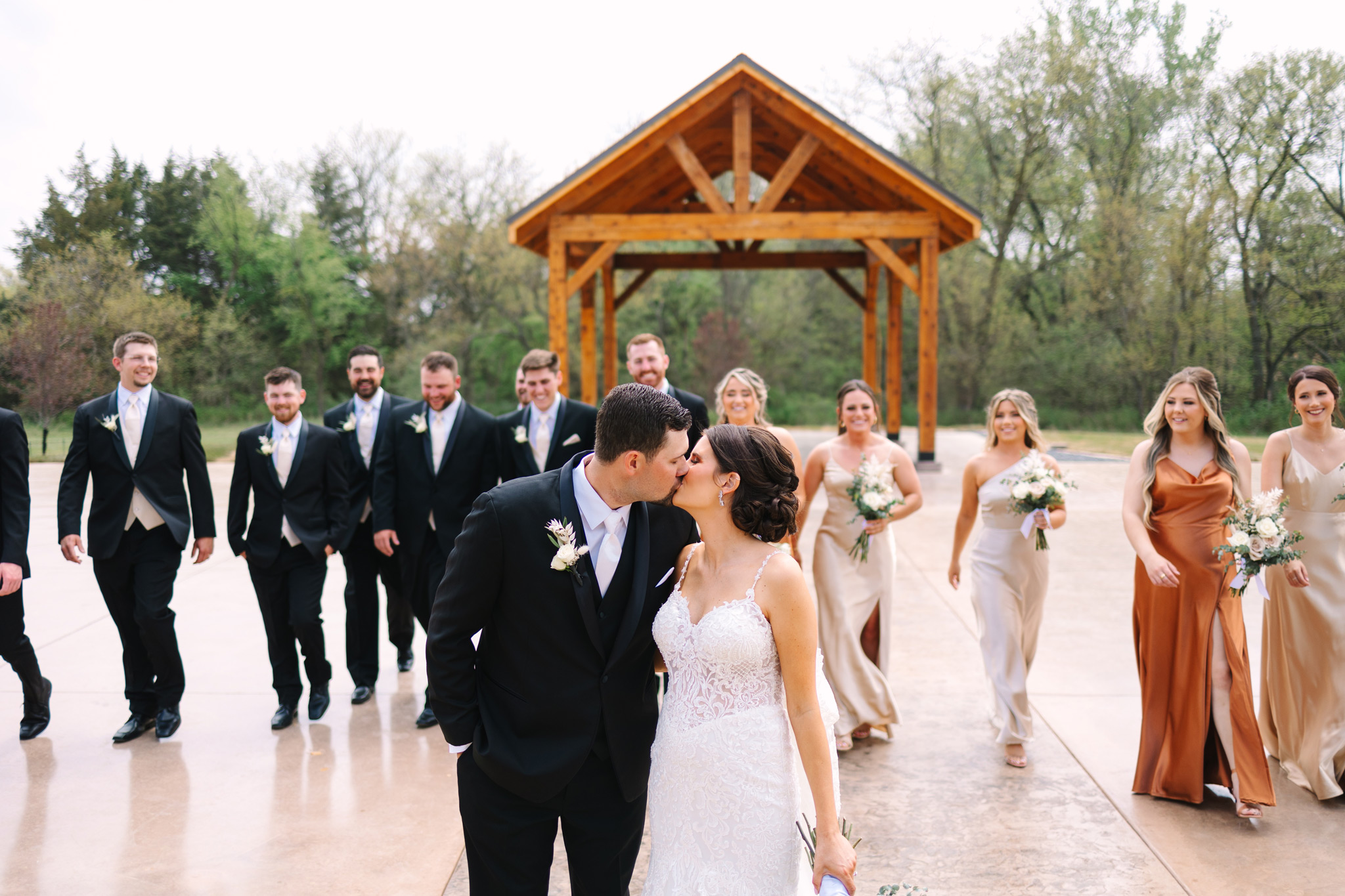 A caucasian bride and groom stop to kiss in front of a wooden pavilion and the wedding party.
