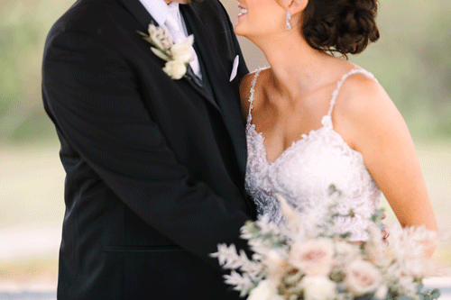 Groom in a black tuxexo on the left side holds his bride who looks up to him while holding a neutral colored bouquet.